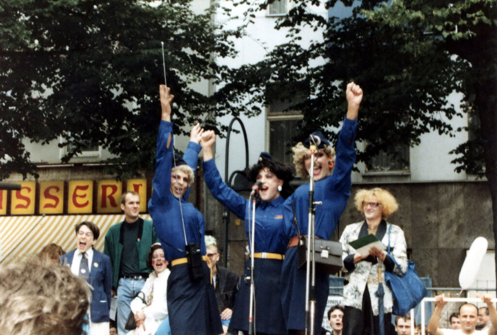 CSD-Bühne 1988, drei Menschen in FJD-Bluse grüßen jubelnd die Menge