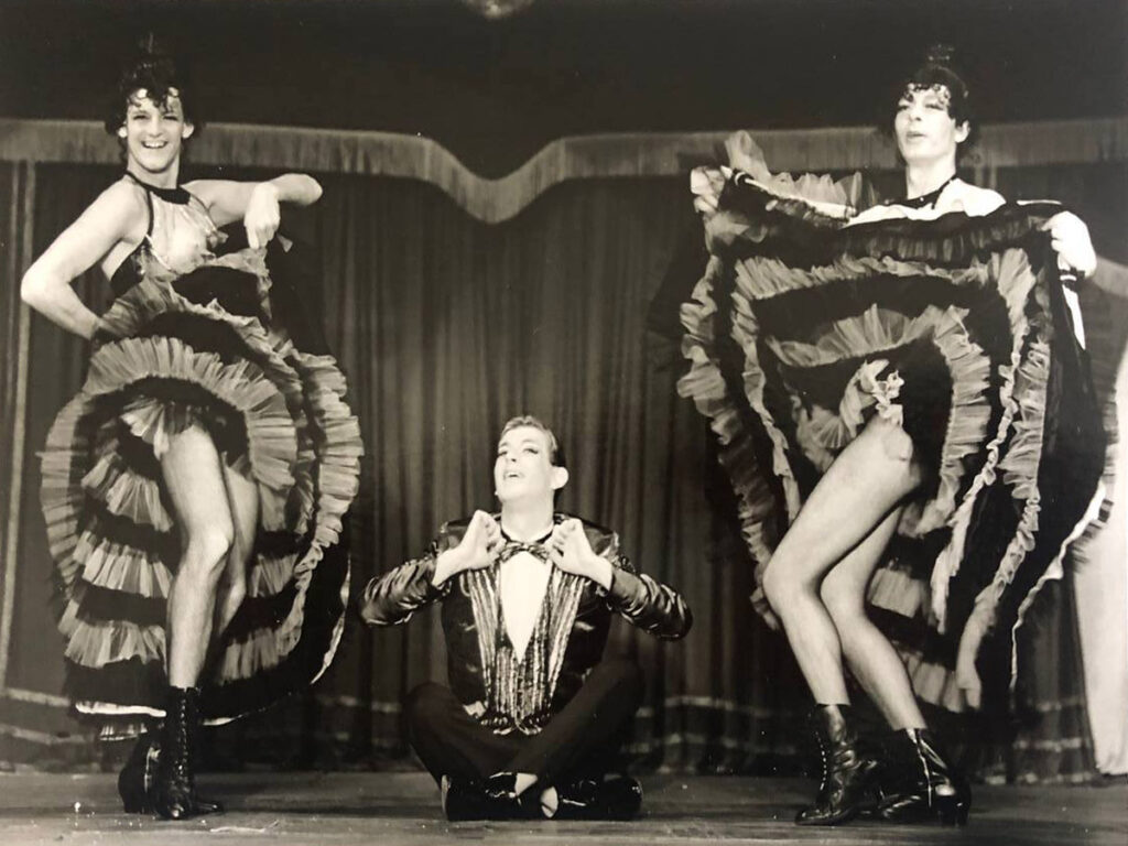 Drei Travestiekünstler tanzen Cancan, einer im Anzug