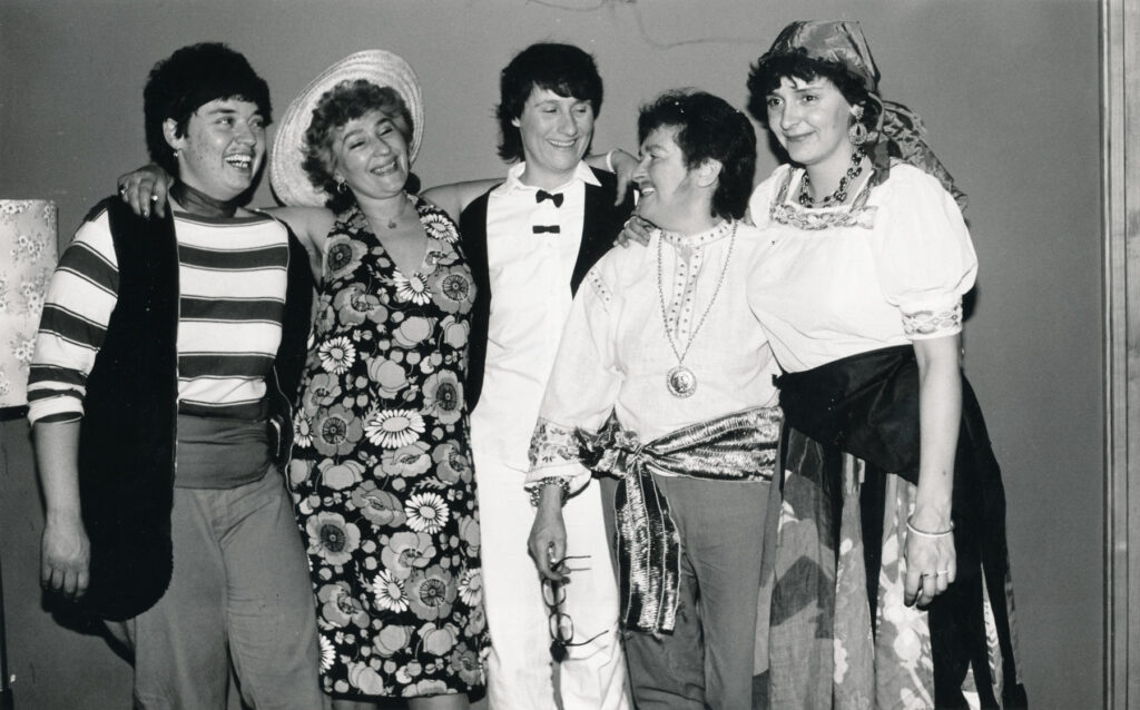 Herrenimitatoren in der DDR - Tommy 2. von rechts - Faschingsfest im Lichtenberger Krug - 1970er Jahre Fünf kostümierte Frauen (Lesben) auf einem Faschingsfest in der Ost-Berliner Kneipe „Lichtenberger Krug“ stehen Arm in Arm in einer Reihe und lachen sich an. Zwei tragen Kleider, drei Frauen tragen Männerkostüme mit Hemden und Westen. Zwei von ihnen haben einen aufgeklebten oder angemalten Bart, eine davon ist Tommy. Quelle des Fotos: FFBIZ-das fem. Archiv / Fotosammlung Rita Thomas - C Rep. 40 Acc. 4 Nr. 174 Berlin-Ost 23