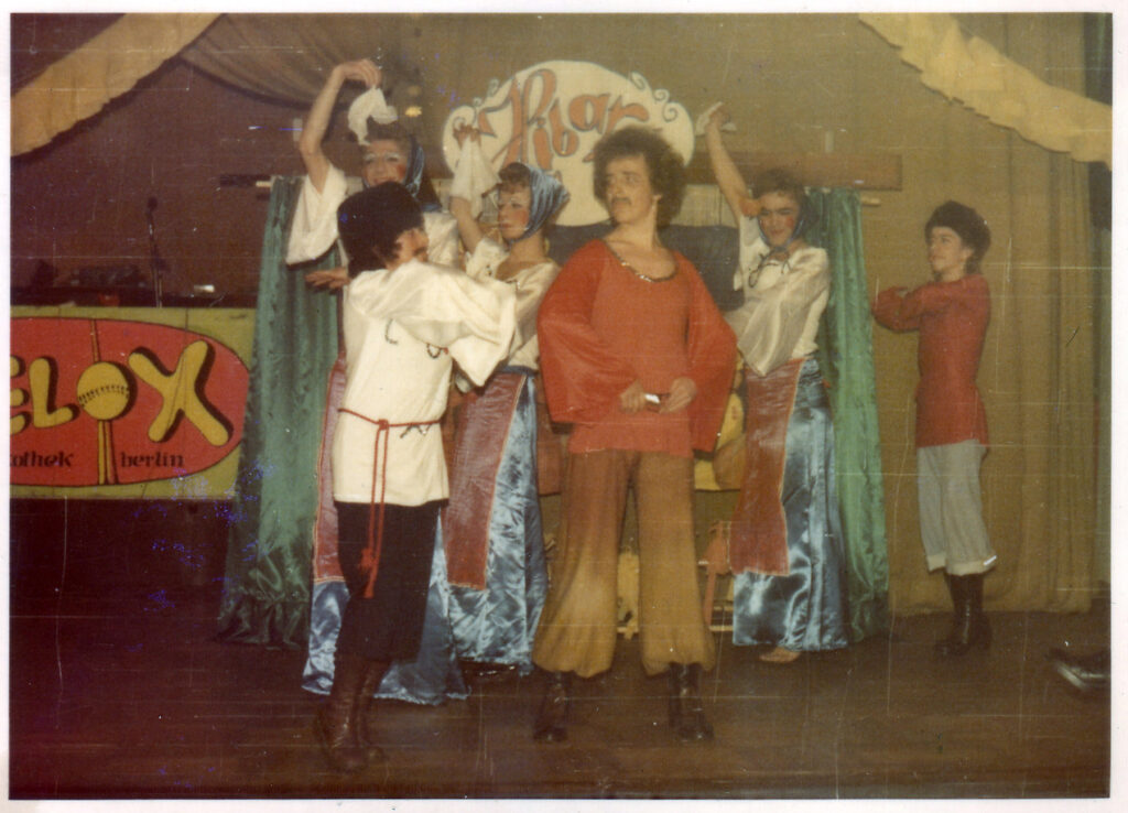Herrenimitatoren in der DDR - 1975 Fasching Bärenschenke - Auftritt Hibaré - Tommy vorne links Eine Faschingsparty 1975 der HIB in der Bärenschenke, auf der Bühne sind 6 Personen zu sehen, drei tragen russische männliche Kosakenkostüme, drei tragen seidig glänzende lange Röcke, Blusen und Tücher im Haar und sind geschminkt. Tommy ist vorne links und trägt einen angeklebten Bart, ein weißes Hemd mit einer roten Kordel als Gürtel und eine schwarze Hose die in hohen Stiefel endet. Tommy macht Tanzsprünge. Im Hintergrund der Bühne ist ein Schild mit dem Schriftzug Hibaré.
