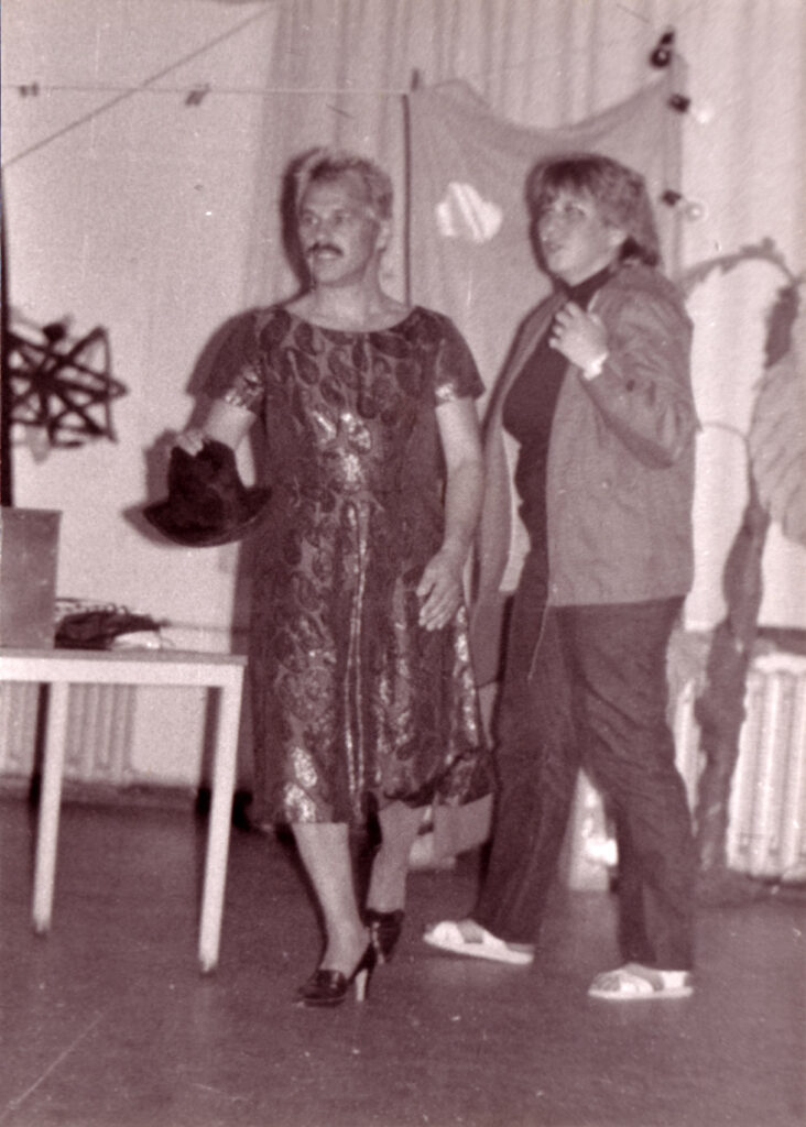 Hochformat – Comedy-Auftritt: Mann mit Schnurbart und Herrenhut in der Hand trägt ein glänzendes Kleid und hochhackige Schuhe und steht neben einer Frau in Jeans, Trainingsjacke und Sandalen vor einer Stofffahne mit einer Glühlampenkette im Hintergrund.