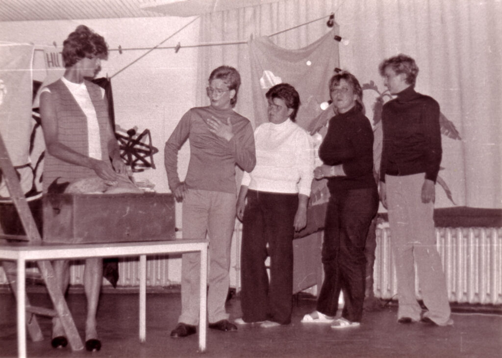 Auf der Bühne des Hibaré-Kabaretts steht ein Tisch mit einem Koffer drauf. Aus dem Koffer holt eine Person in Kleid und hochhakigen Schuhen ein Kleidungsstück raus, neben dem Tisch stehen vier Frauen der Gruppe und interagieren mit Gesten mit der Person hinter dem Tisch.