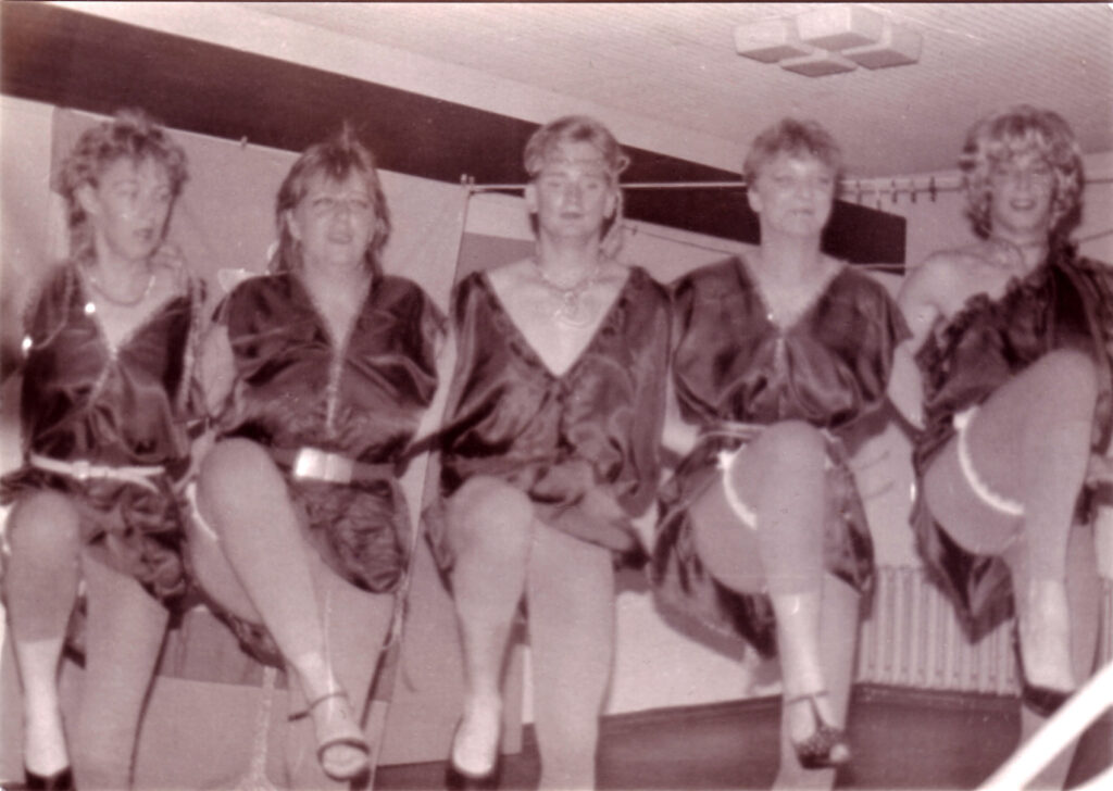 5 Personen haben sich an den Armen eingeharkt und tanzen synchron in einer Reihe – alle tragen dieselben Kostüme: Kleider in glänzendem Stoff und Leggins.