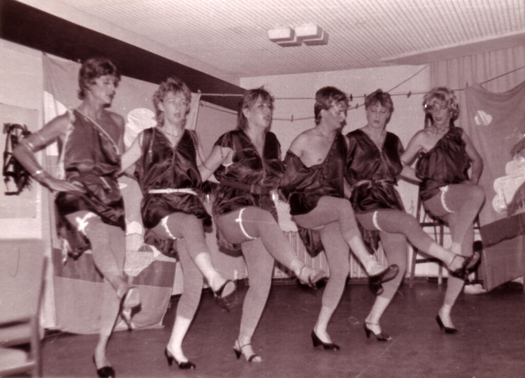 6 Personen haben sich an den Armen eingeharkt und tanzen synchron in einer Reihe – alle tragen dieselben Kostüme: Kleider in glänzendem Stoff und Leggins. Alle tragen Strumpfbänder.