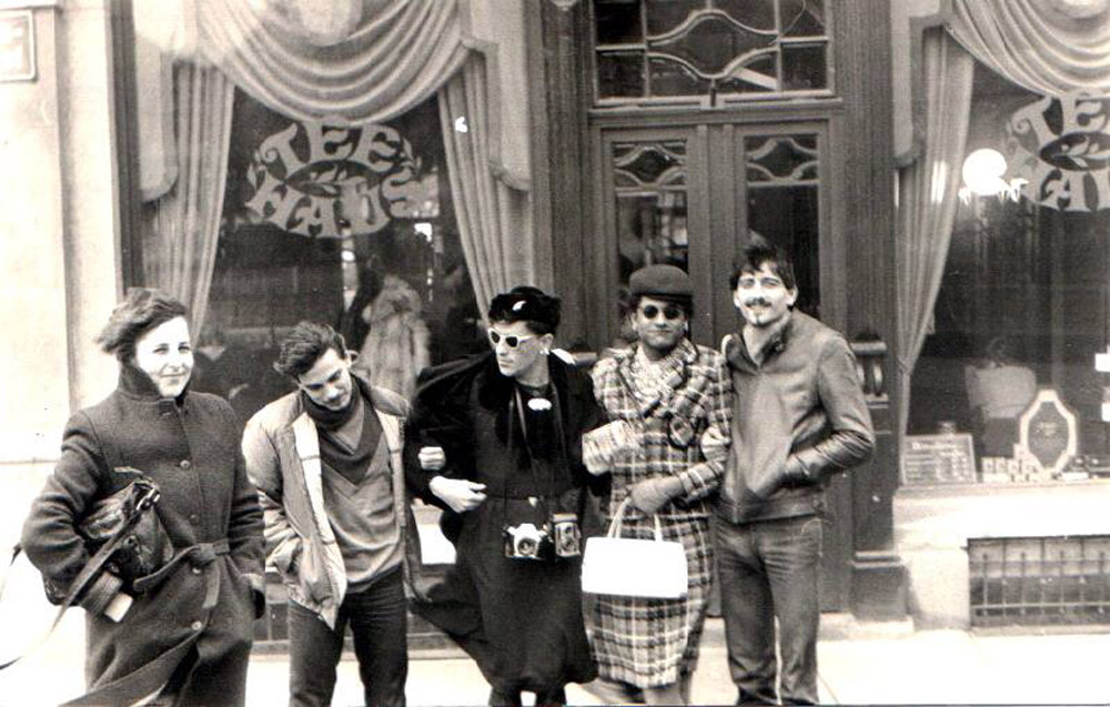 Fünf Personen, davon zwei Männer in Kleidern stehen untergehakt vor einem Teehaus