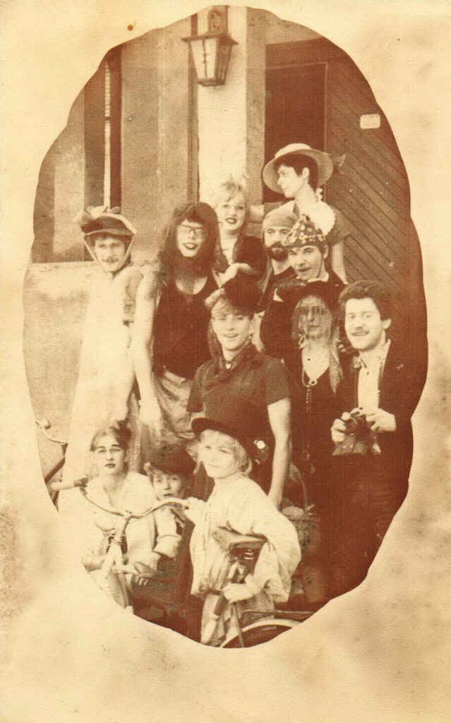 12 Menschen stehen lachend und kostümiert auf der Treppe vor einem Gasthof, im Vordergrund steht ein Fahrrad.